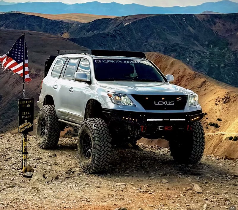 Jason's Lexus LX 570 on 40-inch tires on a high-desert mountain trail, American flag beside it and Rocky Mountain peaks behind